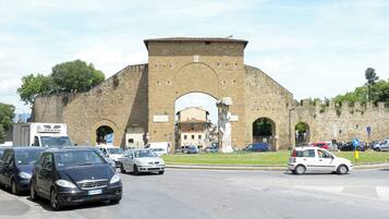 Un arco de piedra histórica con una estatua en primer plano, rodeado de coches aparcados y un cielo despejado.