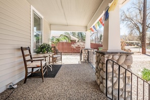 A covered porch with two wooden chairs and a table, a stone wall with a metal railing, and a view of a fenced yard and trees. - Mt Washington House (Colorado Springs)