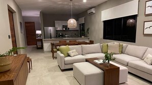 A modern living room with a sectional sofa, ottoman, and a wooden coffee table. There is a dining area with a wooden table and chairs, and a kitchen in the background.