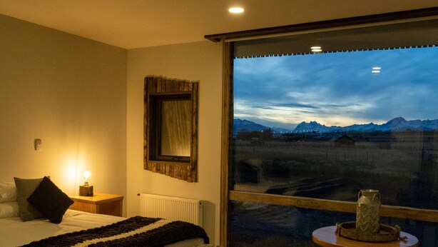 Een slaapkamer met een groot raam, een bed, een nachtkastje met een lamp, een houten bankje en een kleine tafel met een vaas.