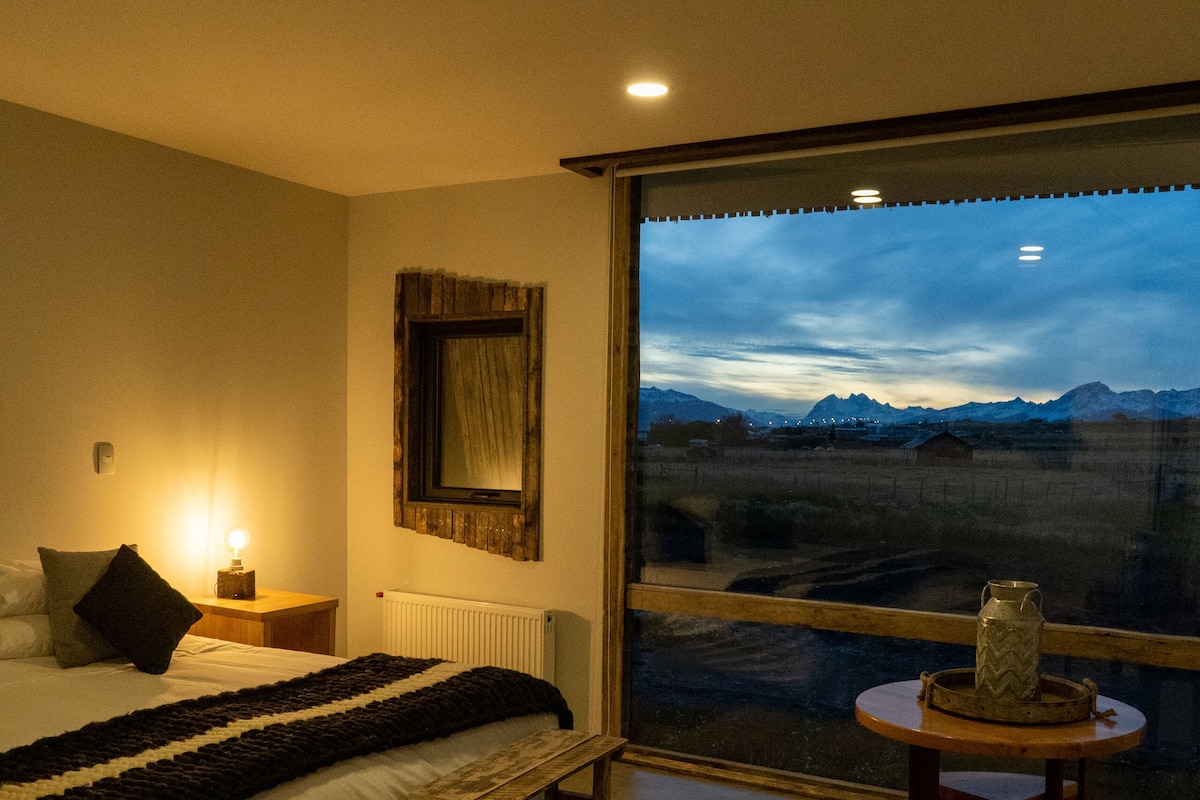 Een slaapkamer met een groot raam, een bed, een nachtkastje met een lamp, een houten bankje en een kleine tafel met een vaas.