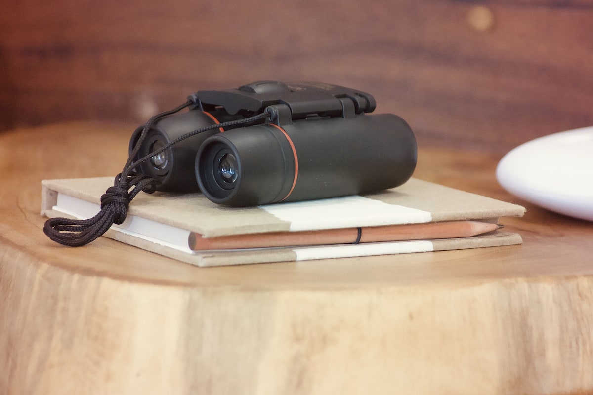 A camera with a strap on a wooden surface, next to a notebook and a pencil.