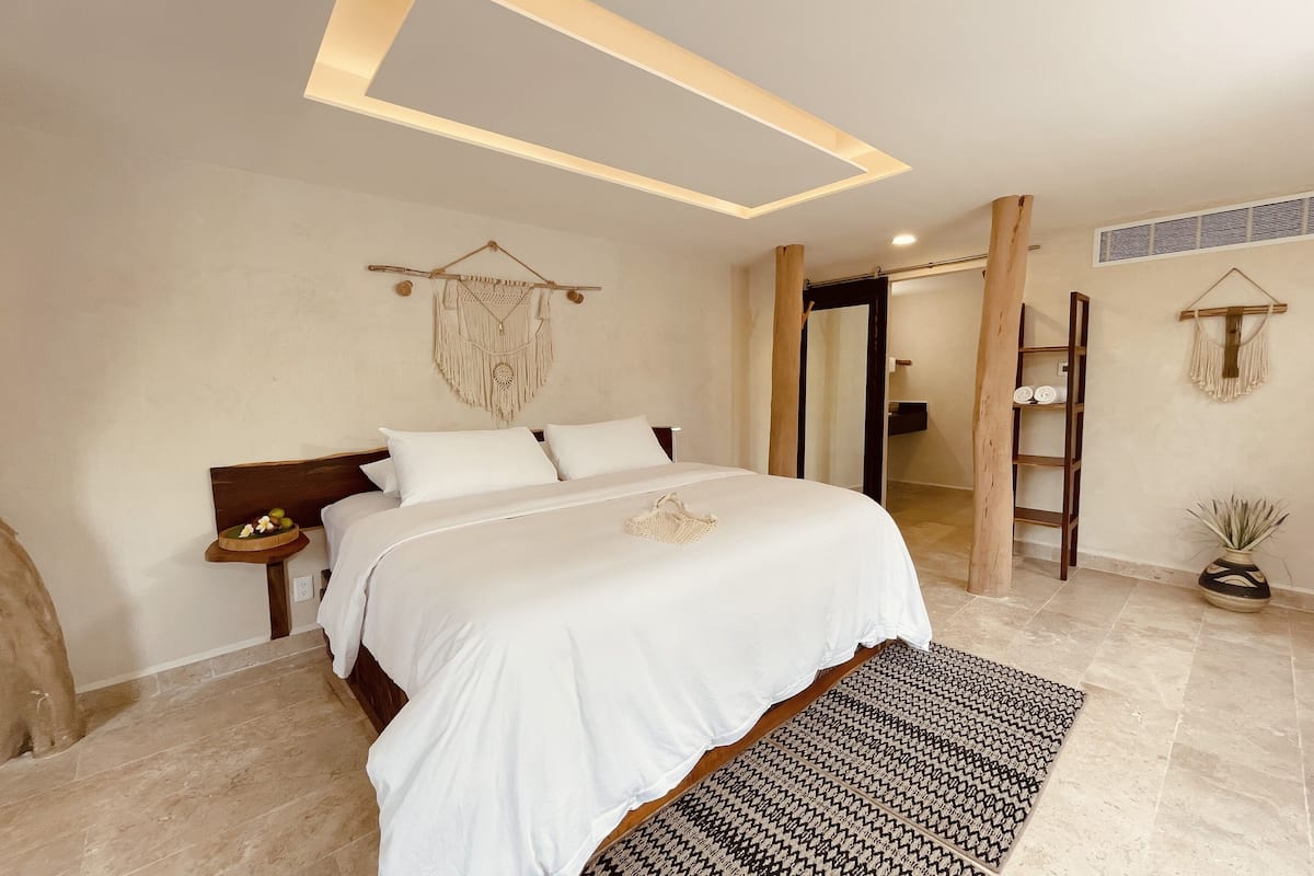 A well-lit bedroom with a large bed, a bedside table, a wall-mounted macrame decoration, and a wooden ladder.