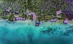 Vista aérea de un resort tropical con bungalows sobre el agua, un muelle y exuberante vegetación.