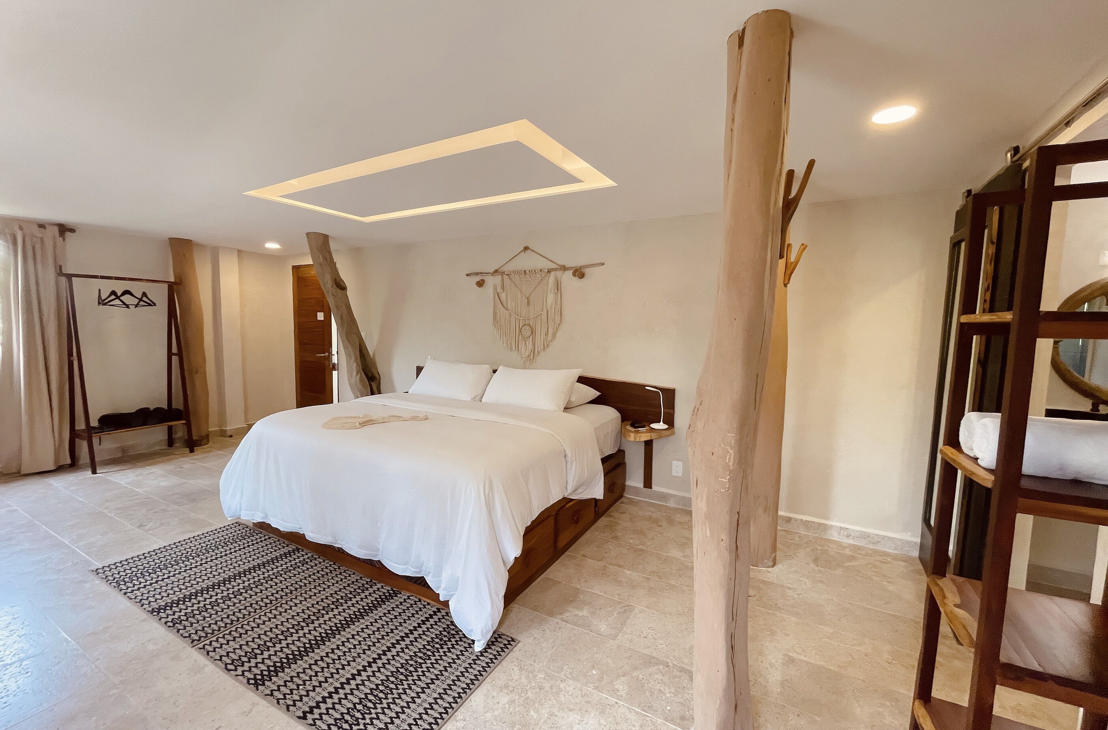 A bedroom with a bed, a wooden headboard, a bedside table, a wall-mounted macrame decoration, and a wooden ladder against the wall.