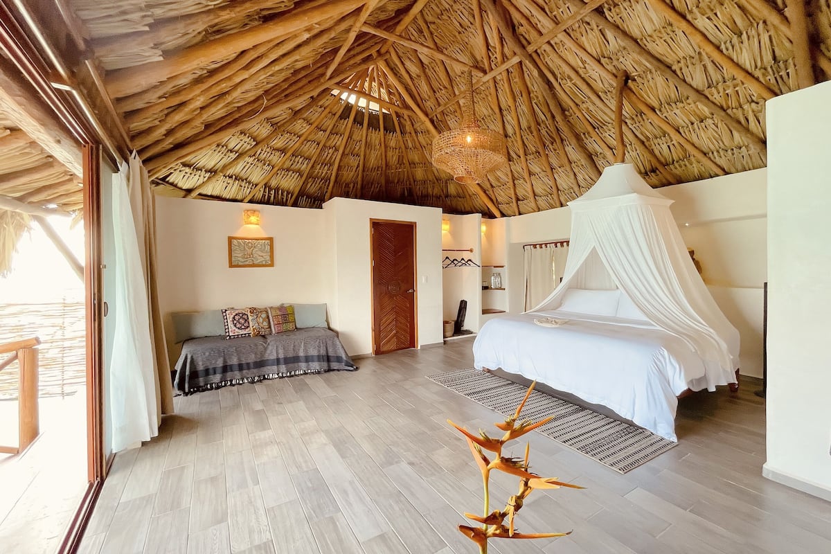 A bedroom with a thatched roof, a bed with a canopy, and a view of the outdoors.