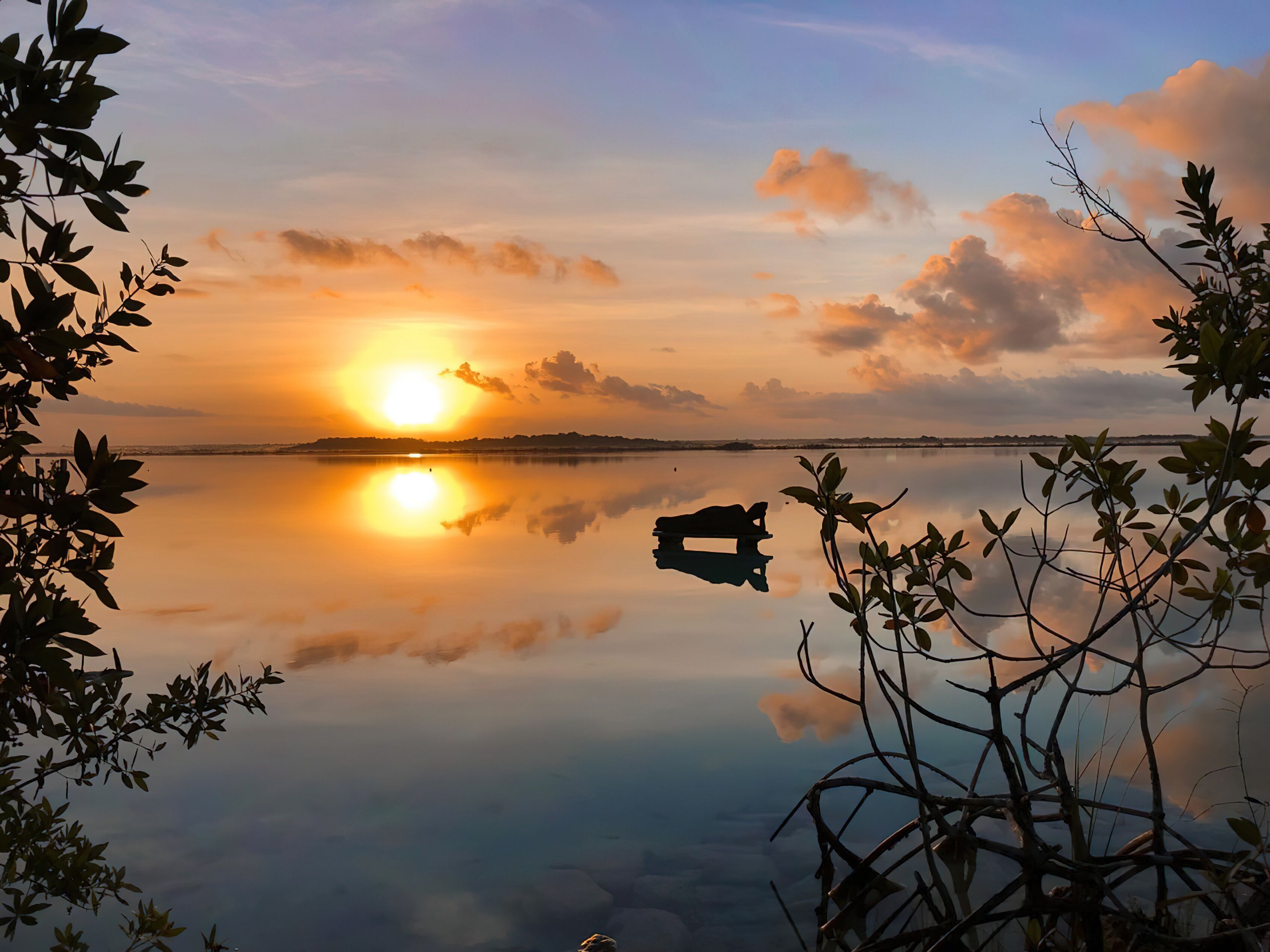 A tranquil sunset over a calm lake with a silhouette of a boat and surrounding foliage.