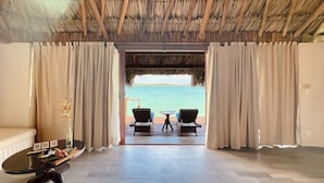 A room with a view of the sea, a thatched roof, a sofa, a table with a vase, and two chairs facing the sea.