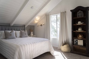 Quarto com uma cama grande, cabeceira de madeira, janela com cortinas, mesa de cabeceira com um vaso e um armário de madeira com chapéus e micro-ondas.