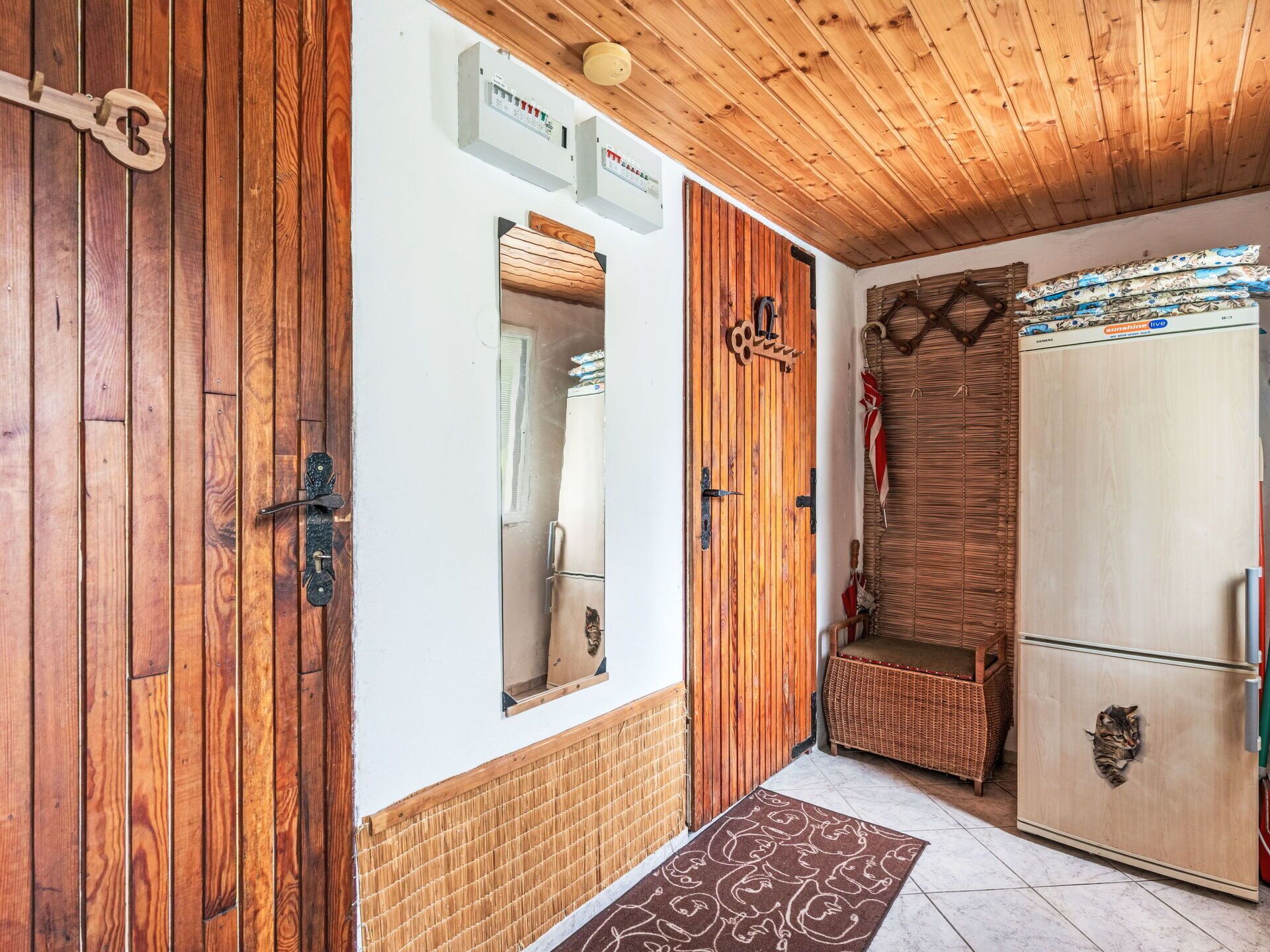 Una habitación con paredes de madera, suelo de baldosas, techo de madera, un refrigerador, una silla de mimbre y una puerta con mirilla.