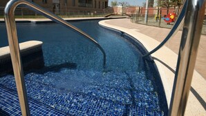 A swimming pool with a tiled floor, metal handrails, and a view of a multi-story building in the background. - Beautiful apartment for 4 people with WIFI, pool, TV, terrace and pets allowed (Empuriabrava)