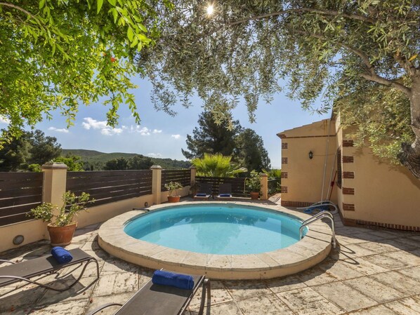 Ein runder Pool mit einer Metallleiter, umgeben von einer gefliesten Terrasse. Liegestühle und Kübelpflanzen sind vorhanden.