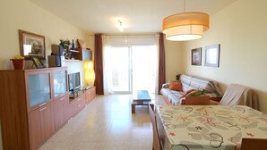 A well-lit living room with a sofa, a TV on a wooden cabinet, and a dining area with a table. - Beautiful apartment for 4 people with pool, A/C, TV and terrace (Sant Carles De La Ràpita)