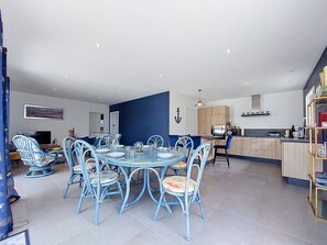 A modern kitchen with wooden cabinets, a dining area with a round table and blue chairs, and a living area with a sofa and TV.