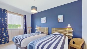 Une chambre avec deux lits, une chaise, une fenêtre avec des rideaux et des œuvres d’art accrochées au mur.