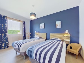 A bedroom with two beds, a chair, a window with curtains, and artwork on the wall.