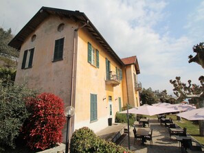 A two-story building with a balcony, outdoor seating area with umbrellas, and a garden with red plants. - Lovely apartment for 3 guests with WIFI, TV and balcony (Ghiffa)