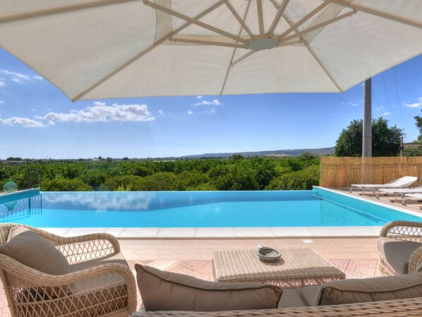 A swimming pool with wicker furniture and an umbrella, surrounded by greenery and a clear sky.
