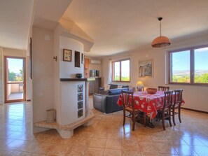 A spacious living area with a dining table, chairs, and a sofa. There is a kitchen area with a microwave and a television. Large windows provide natural light and views of the outdoors.