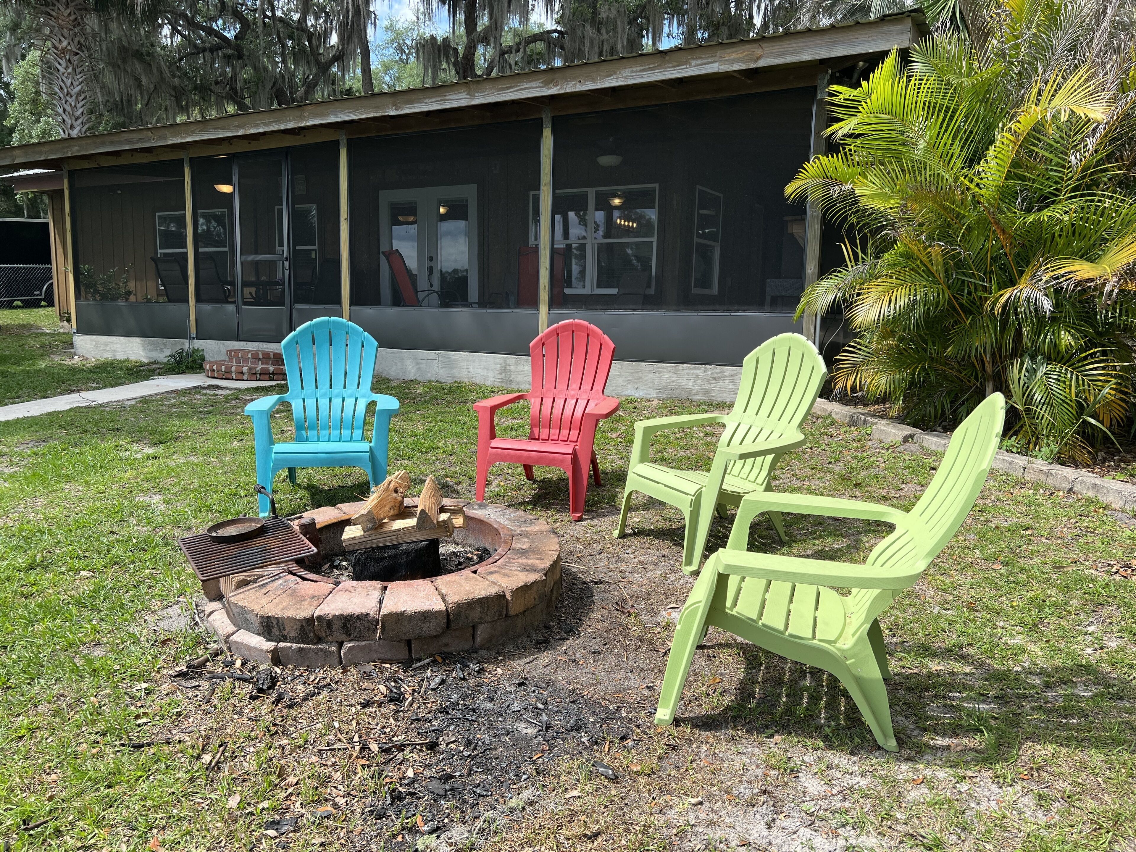 A fire pit with three Adirondack chairs in blue, red, and green colors, positioned in front of a building with large windows and a covered patio area.