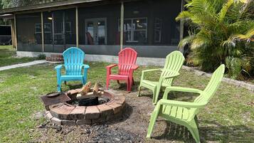 A fire pit with three Adirondack chairs in blue, red, and green colors, positioned in front of a building with large windows and a covered patio area.