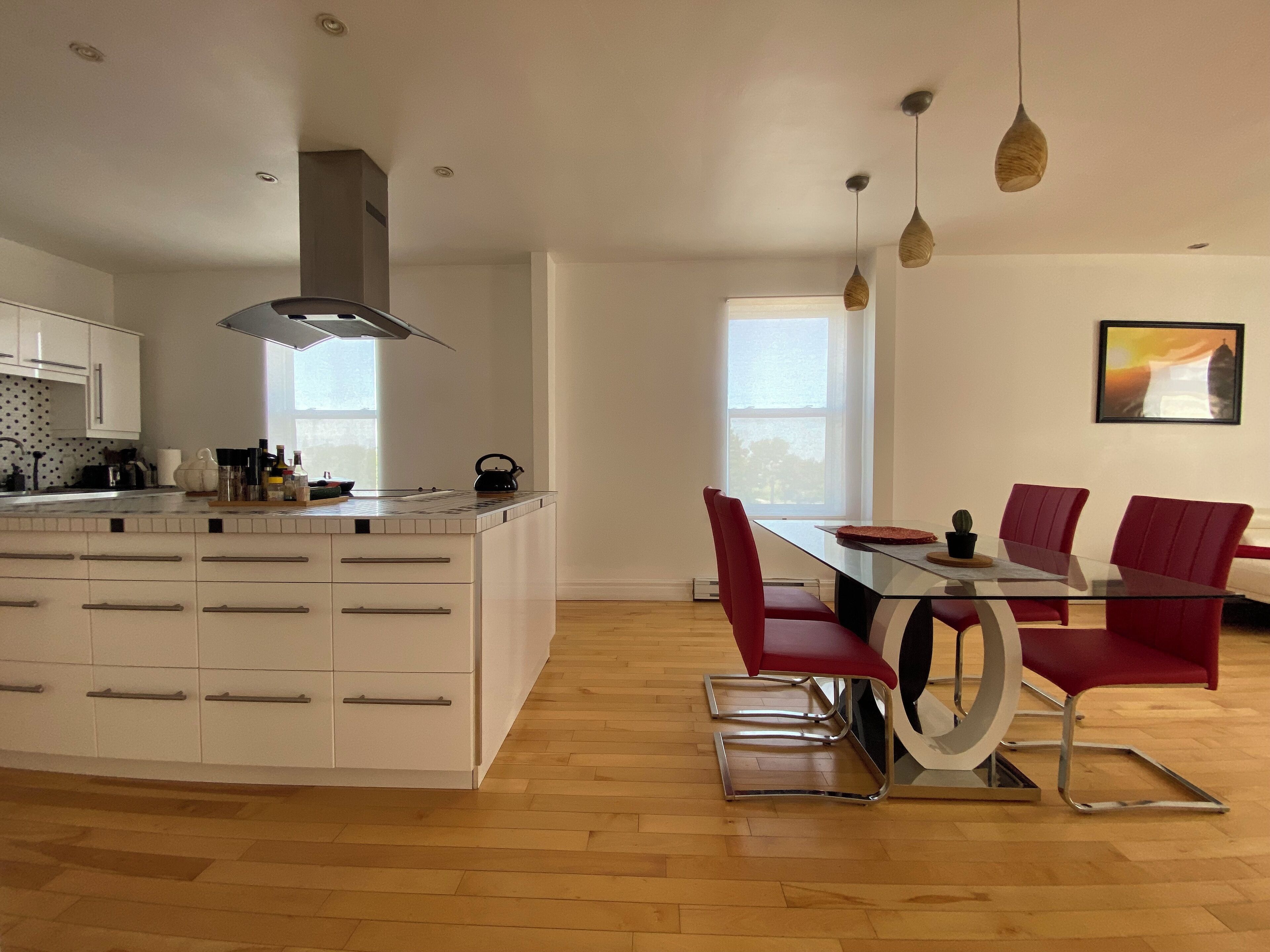 Une cuisine moderne avec des meubles blancs, un grand îlot central et un coin repas agrémenté de chaises rouges et d’une table en verre.