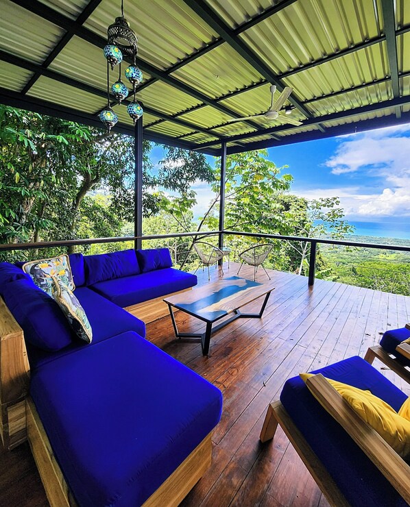Un espacio de estar techado al aire libre con sofás azules y una mesa de centro de madera, con vistas a un paisaje exuberante.