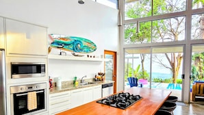 Une cuisine moderne dotée d’un îlot central en bois, d’appareils électroménagers en acier inoxydable et de grandes fenêtres offrant une vue sur l’extérieur.