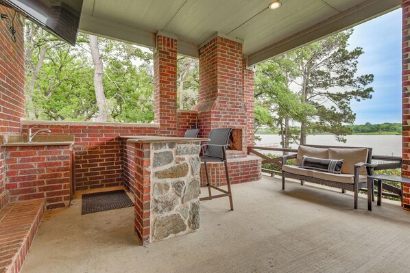 A covered outdoor area with a brick barbecue, stone steps, a cushioned bench, and a view of trees and water.