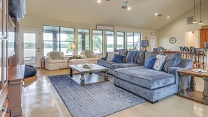 A spacious living room with a large grey sectional sofa, a coffee table, and a ceiling fan. There are large windows providing a view of the outdoors. - Dock & Game Room: Alvarado Lake Getaway! (Alvarado)