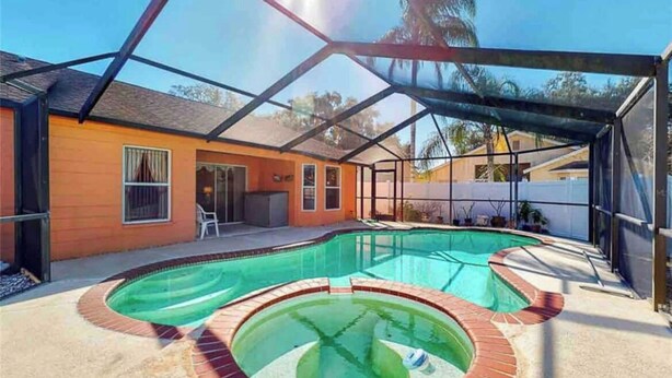 A swimming pool with a glass enclosure, a patio area, and a house with a red exterior. - Relaxing 3 Bedroom Pool Home (Tampa)