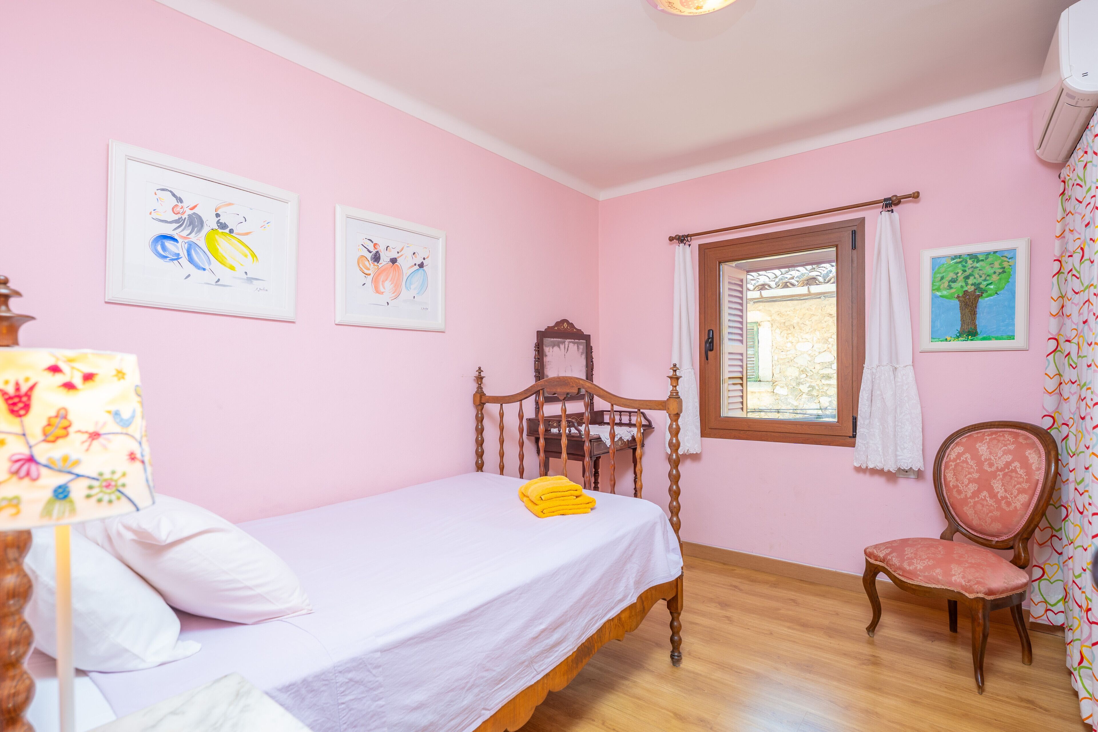 Ein Schlafzimmer mit einem Einzelbett, einem Holzhocker, einem Fenster mit Vorhängen und zwei gerahmten Bildern an der Wand.