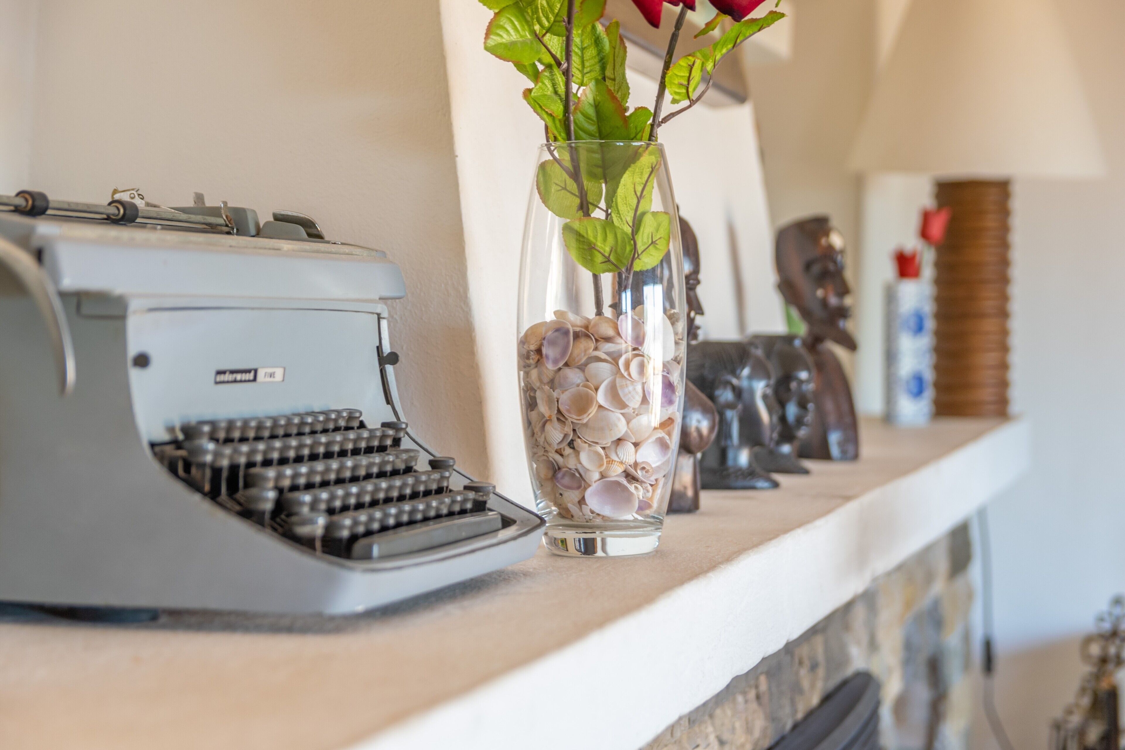 Una máquina de escribir antigua sobre una repisa, un florero con conchas marinas y una flor roja, y una escultura de madera.