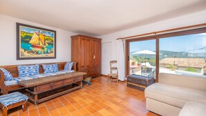 A living room with a wooden sofa, a bench, a cushioned armchair, and a large window with a view of the countryside. - CAN VIDAL - Cozy House with Private Pool and Free WiFi (Campanet)