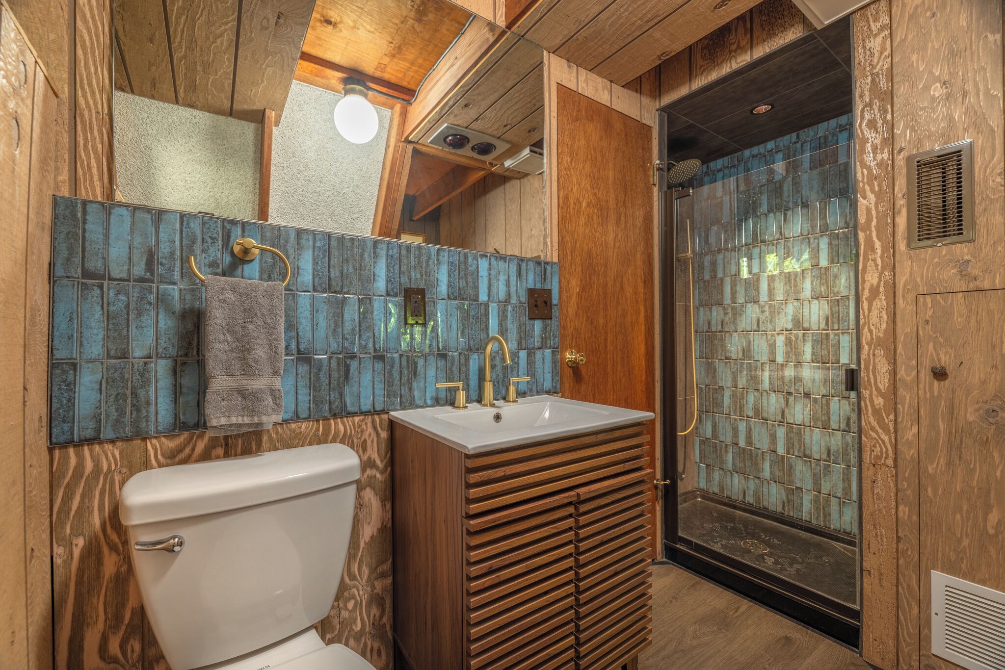 A bathroom with a wooden vanity, a white toilet, and a shower with blue mosaic tiles.