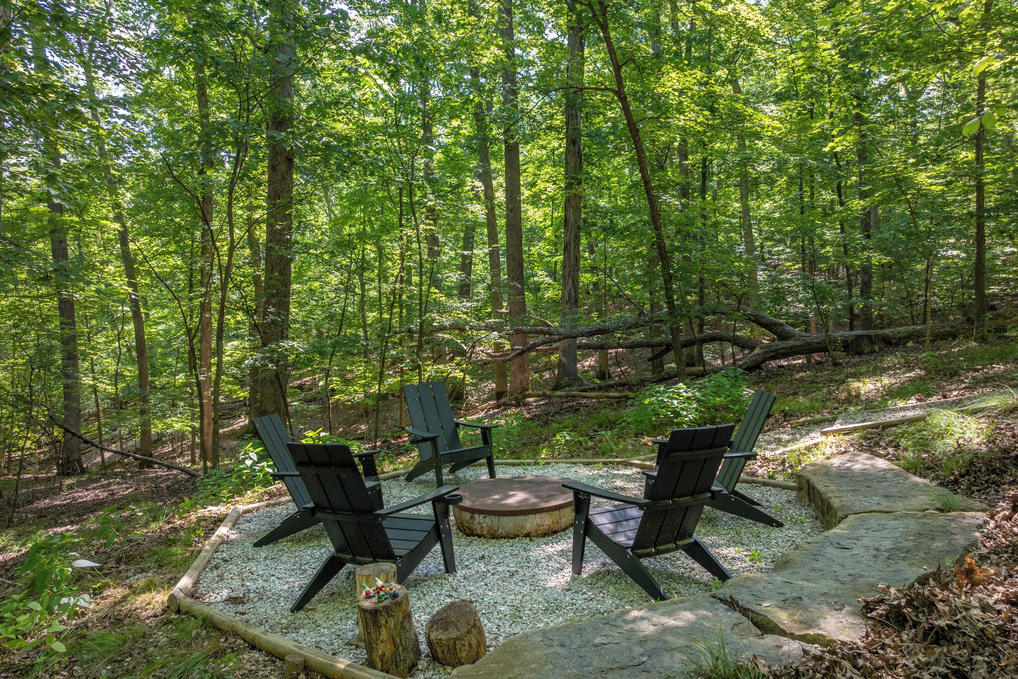 A circular seating area with four chairs and a central fire pit, surrounded by a stone border, set in a forested area.