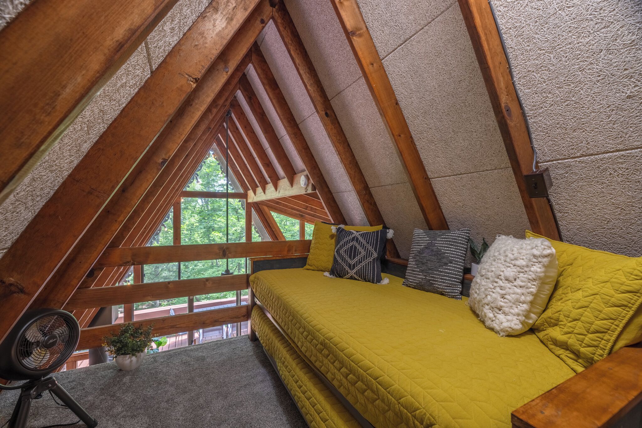 A cozy attic bedroom with a wooden ceiling, a bed with yellow bedding, and a view of greenery outside.
