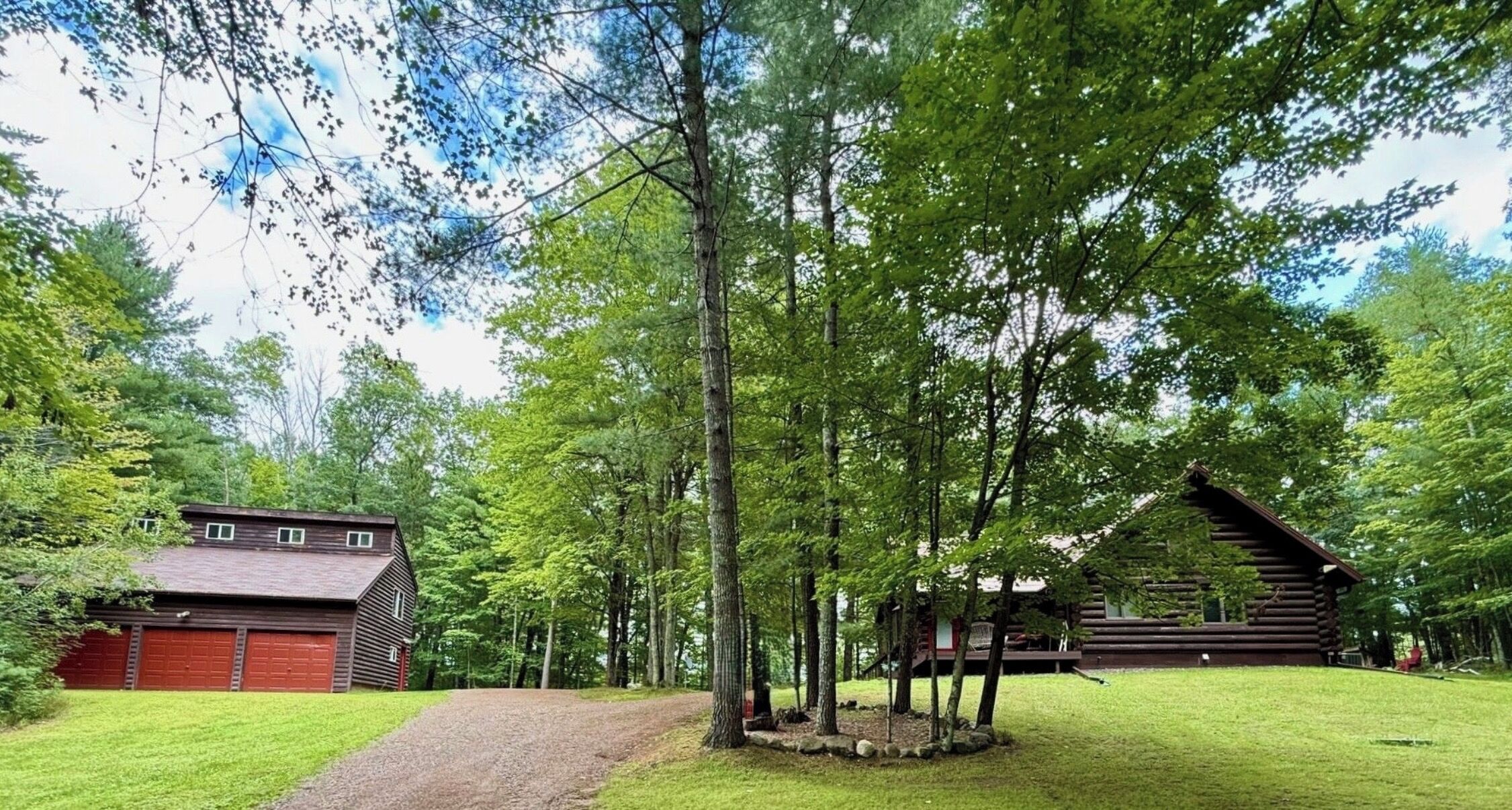 Une cabane rustique aux murs en bois, avec une allée en gravier et une grange rouge, située dans une zone boisée.