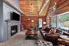 A log cabin interior with a fireplace, leather sofa, patterned armchair, and a view of the outdoors. - Family Log Cabin - Lake, Golf, Pool, Hike, Ski-ATV & Game Room - Northwoods WI (Danbury)