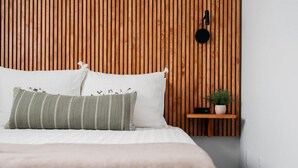 Una cama con ropa de cama blanca y un almohadón a rayas, un pequeño estante de madera con una planta, y un altavoz montado en la pared.