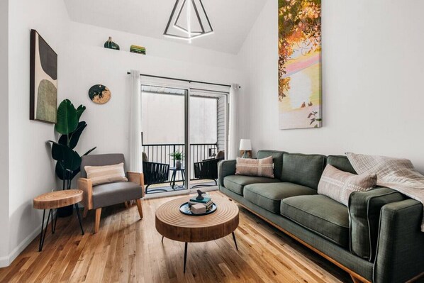 Un salon moderne avec un canapé vert, un fauteuil gris, une table basse en bois et une grande fenêtre donnant sur l’extérieur.