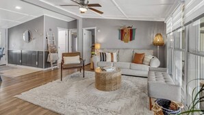 Un salon spacieux avec un canapé gris, un fauteuil en cuir marron, une table basse ronde et un ventilateur de plafond.