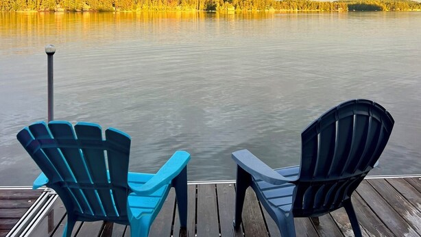 Ett trädäck med två Adirondack-stolar som vetter mot en stilla sjö med en skogbeklädd strand.