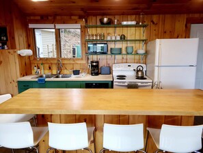 A wooden cabin kitchen with a central island, white chairs, a stove, a sink, and shelves above the counter. - Private Lake House with Docks & Views for Days (Shapleigh)