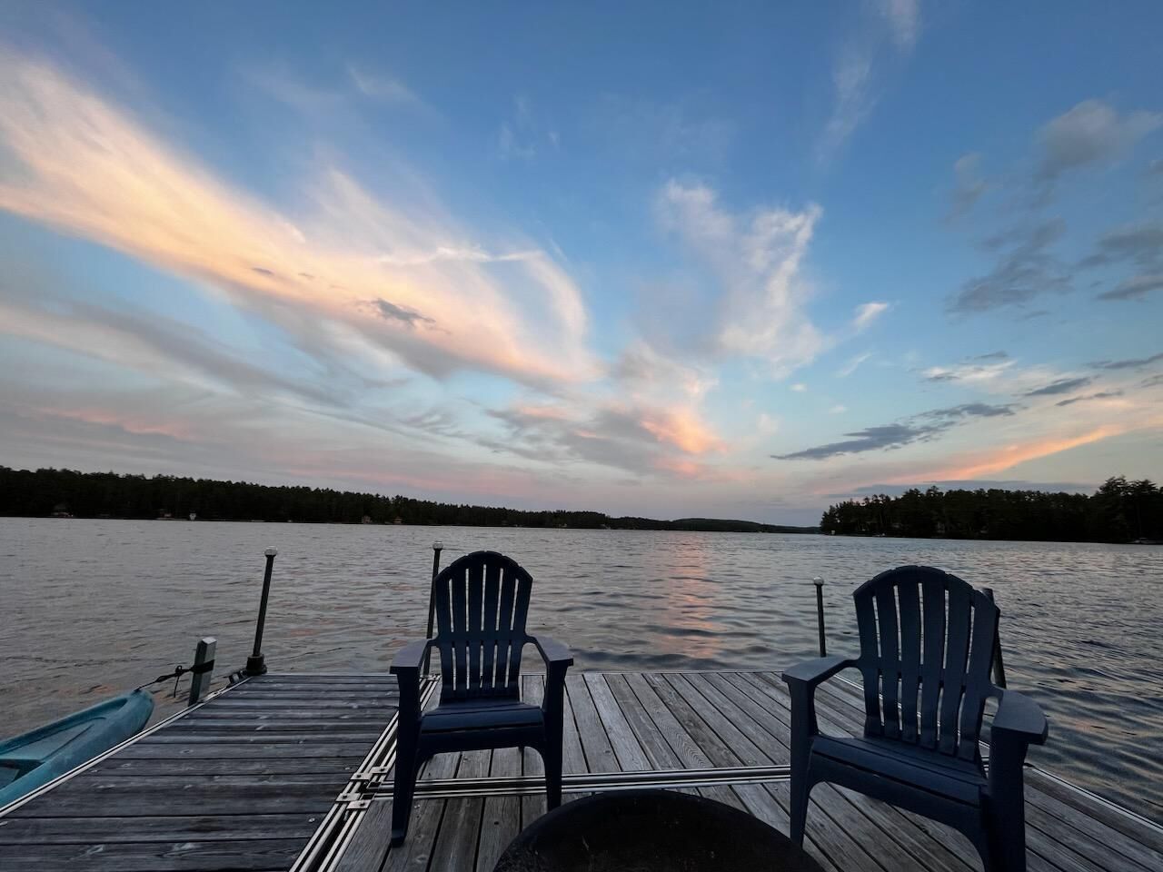 A wooden dock with two Adirondack chairs facing a calm lake at sunset.