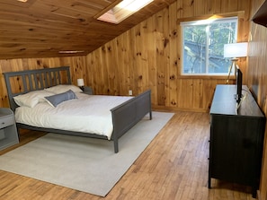 A bedroom with a wooden ceiling, a bed with white bedding, a nightstand, a desk with a lamp, and a window with a view of trees. - Private Lake House with Docks & Views for Days (Shapleigh)