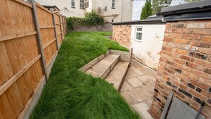 Un petit jardin à l’arrière de la maison, délimité par une clôture en bois et un mur de briques, avec un petit espace verdoyant comprenant de l’herbe et des marches en pierre.