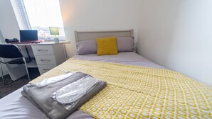 Un lit bien fait, recouvert d’une couverture à motifs jaunes, avec un oreiller gris et un autre oreiller de couleur moutarde. On y voit un bureau avec une chaise et un ordinateur portable.