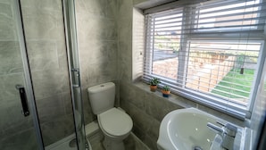 Une salle de bain avec une porte de douche en verre, des toilettes, un lavabo et une fenêtre avec des stores.