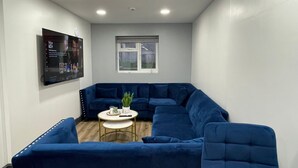 Un salon moderne avec un canapé d’angle bleu, une petite table ronde ornée d’une plante et un téléviseur à écran plat fixé au mur.